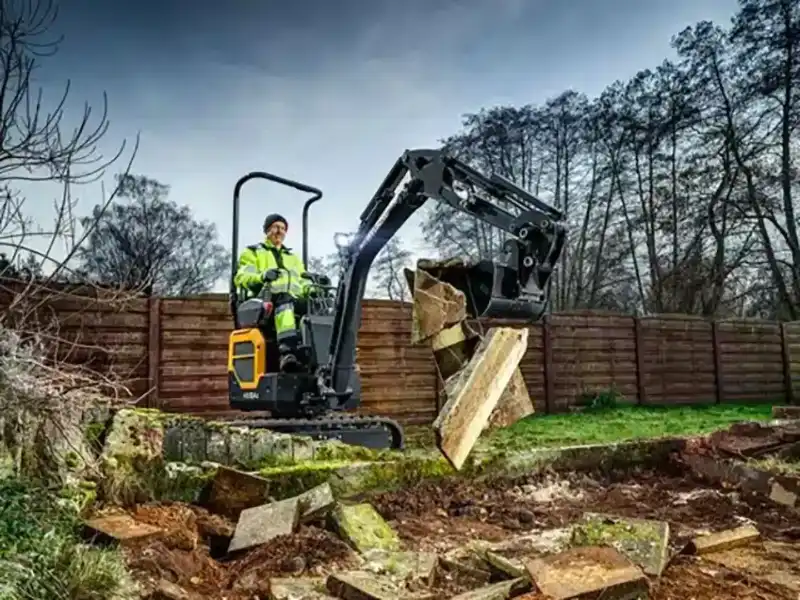 hyundai-hx10a-z-compact-excavator-Compact excavator with operator in high-visibility jacket digging in a yard, logs and soil piles near a wooden fence.
