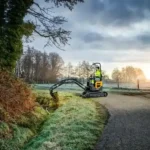 hyundai-hx10a-z-compact-excavator-Yellow compact excavator working on a grassy roadside near trees at sunset, with bucket lowered and tracks on gravel path.