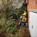 hyundai hx10a z compact excavator Compact excavator with two workers in high-visibility vests digging beside a brick wall and white fence panel outdoors